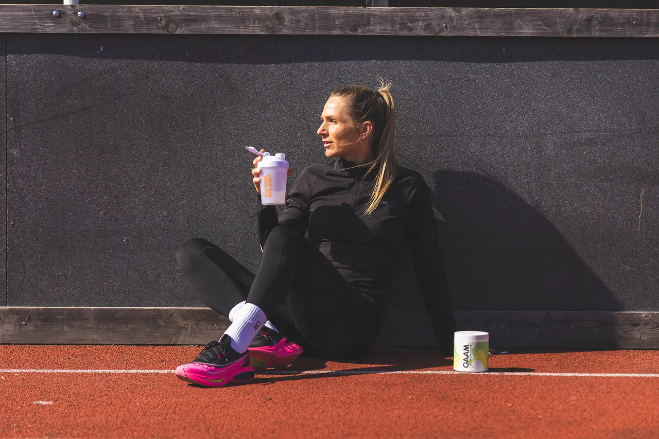 Kvinna i löparträning sitter på friidrottsbana med proteinshaker i handen och GAAM kosttillskott bredvid sig