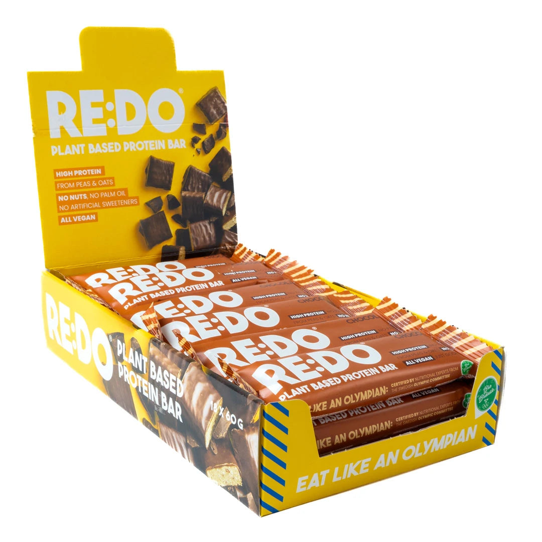 18 x REDO Bar 60 g Chocolate Chock avoimessa keltaisessa esittelylaatikossa täynnä ruskeita proteiinipatukoita.